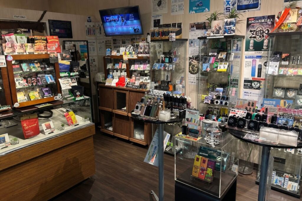 明るく整理された vapeショップ店内。ガラスショーケースや木製カウンターにカラフルな電子タバコ・VAPEデバイス、リキッド、パッケージが豊富に並び、壁には商品ポスターが多数貼られている。奥の壁にテレビが設置され、店内全体が商品で充実している様子。