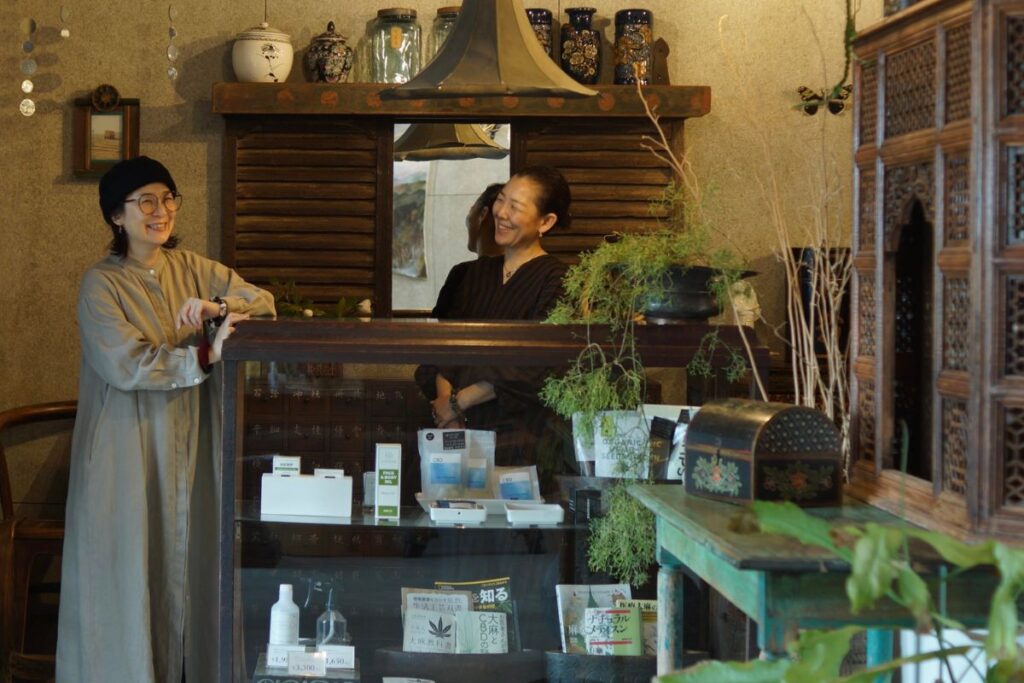 古風な内装のCBDショップで、アンティークのガラスショーケース越しに笑顔で会話する2人の女性。ケース内にはCBD製品と大麻・CBD関連書籍が並ぶ。