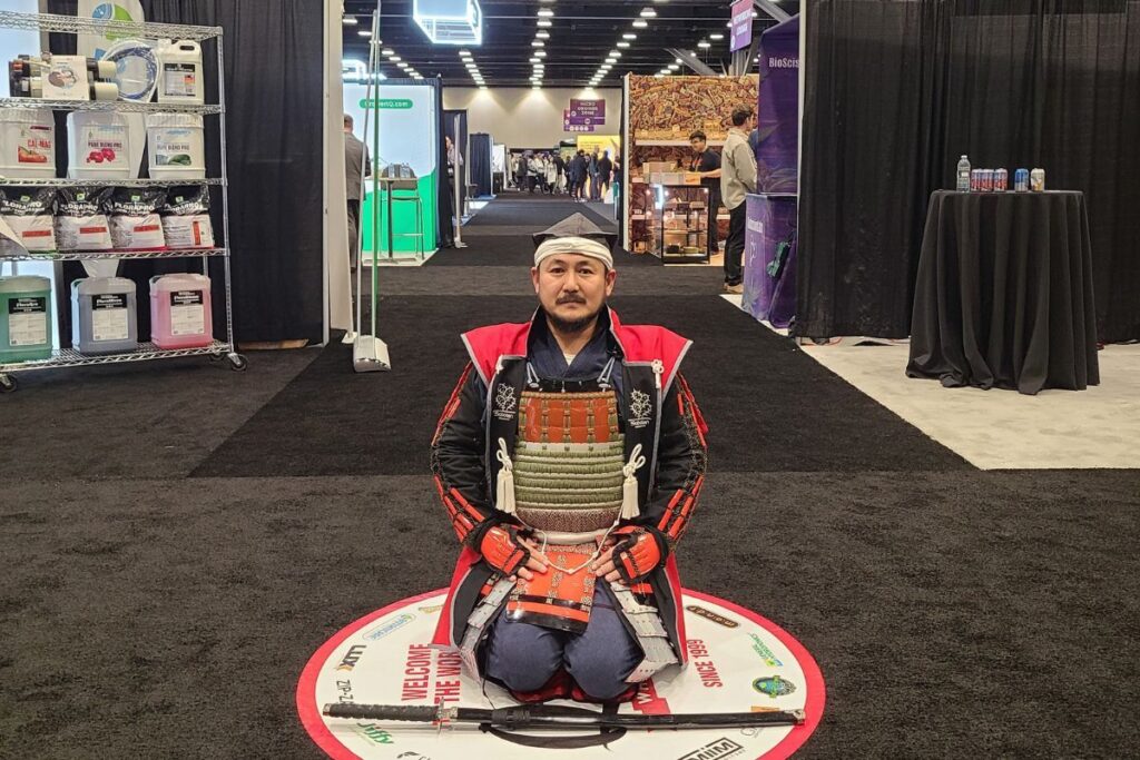 貿易展示会会場で正座する赤と黒のサムライ甲冑姿の日本人男性（刀を前に置いて）