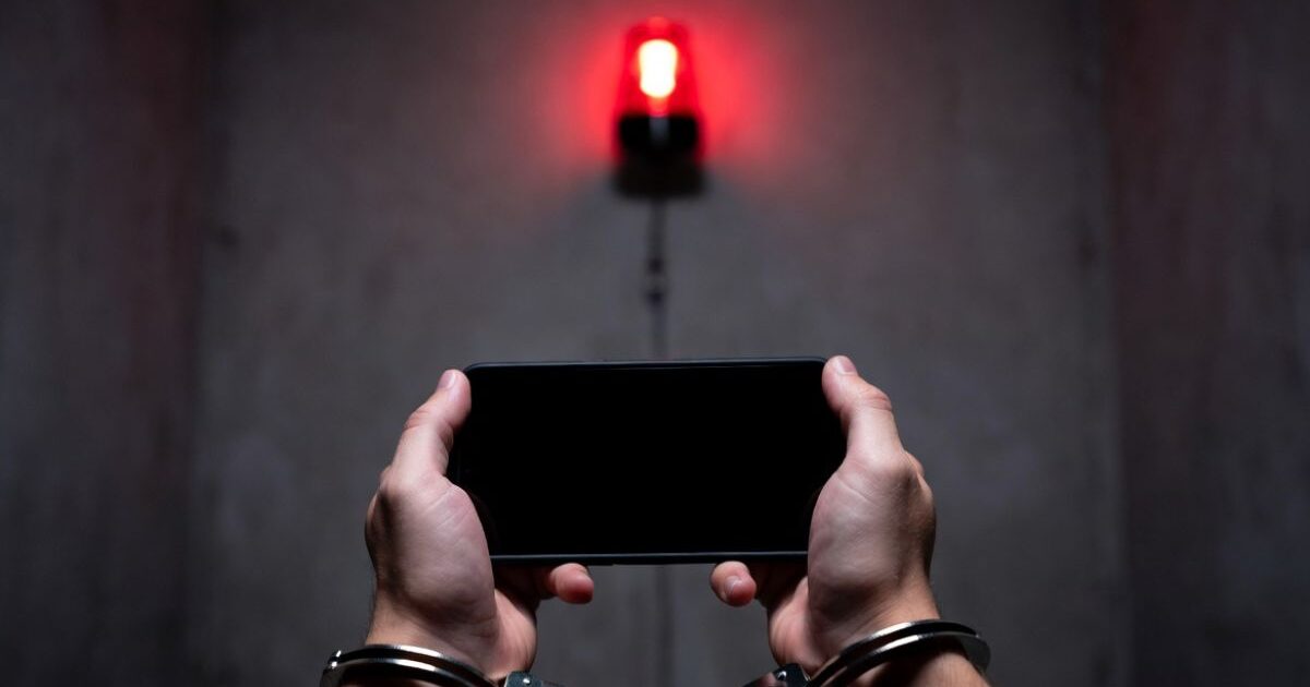 手錠をかけられた手でスマートフォンを持ち、背後に赤い警告灯が光る様子