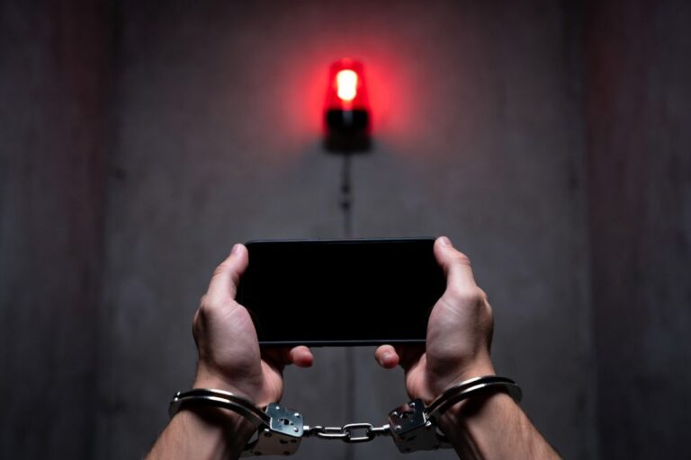 手錠をかけられた手でスマートフォンを持ち、背後に赤い警告灯が光る様子