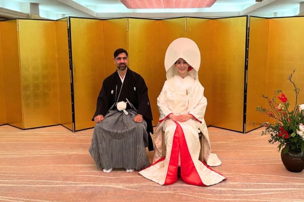 黒の紋付袴を着た男性と、白い打掛に角隠し（つのかくし）を付けた女性が、金色の屏風の前で並んで座っている結婚式の記念写真