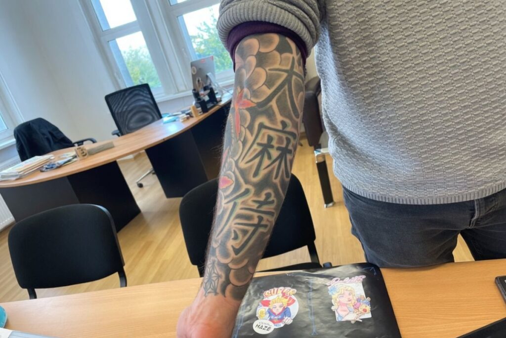 人物の左腕に彫られた大麻の葉と漢字のタトゥー。雲のような背景に赤い花がアクセントで入った、黒と灰色のグラデーションが美しい和彫り風のタトゥーアート。腕を下に伸ばした状態で撮影され、手首近くに小さな蜘蛛の巣のようなデザインも見える。