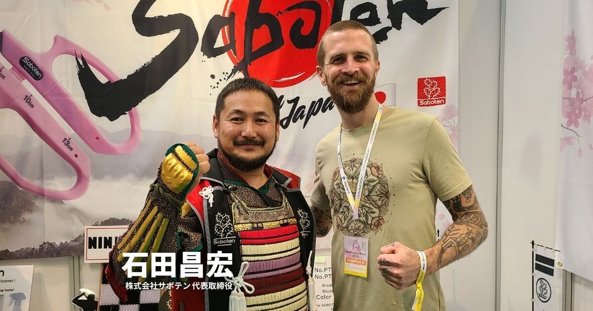 Sabotenブースで記念撮影するサムライ甲冑姿の日本人男性とタトゥー入りの外国人男性