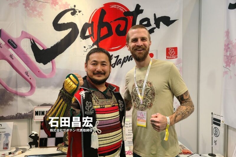 Sabotenブースで記念撮影するサムライ甲冑姿の日本人男性とタトゥー入りの外国人男性