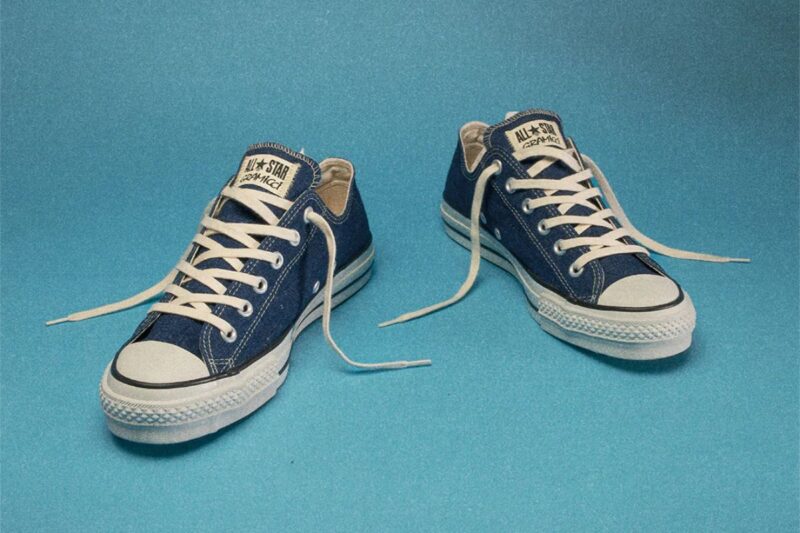 青いデニム生地のコンバース オールスター（Converse All Star）スニーカー。白いレースと白いラバーソールが特徴のクラシックな一足が、明るい青色の背景に並べて置かれている
