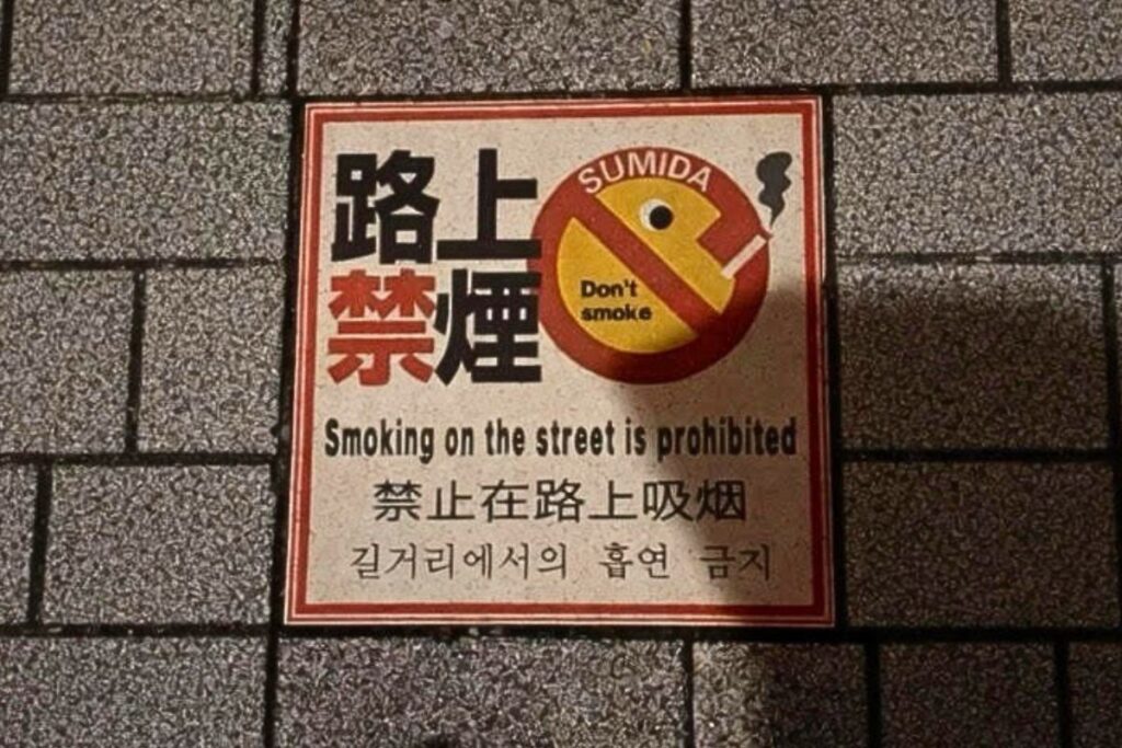 灰色のタイル舗装の上に置かれた赤枠の禁煙看板。中央に禁止マークと『路上禁煙』『Smoking on the street is prohibited』などの多言語表記がある
