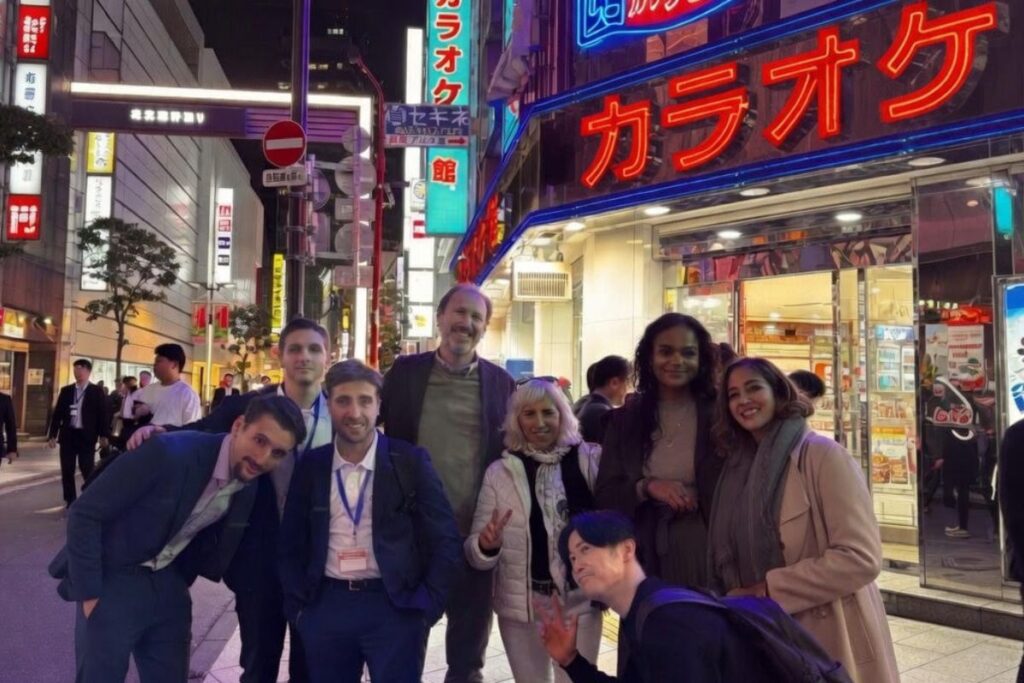 夜の東京の街中で、ネオン輝くカラオケ店の前で笑顔でポーズを取る8人の国際的なグループ