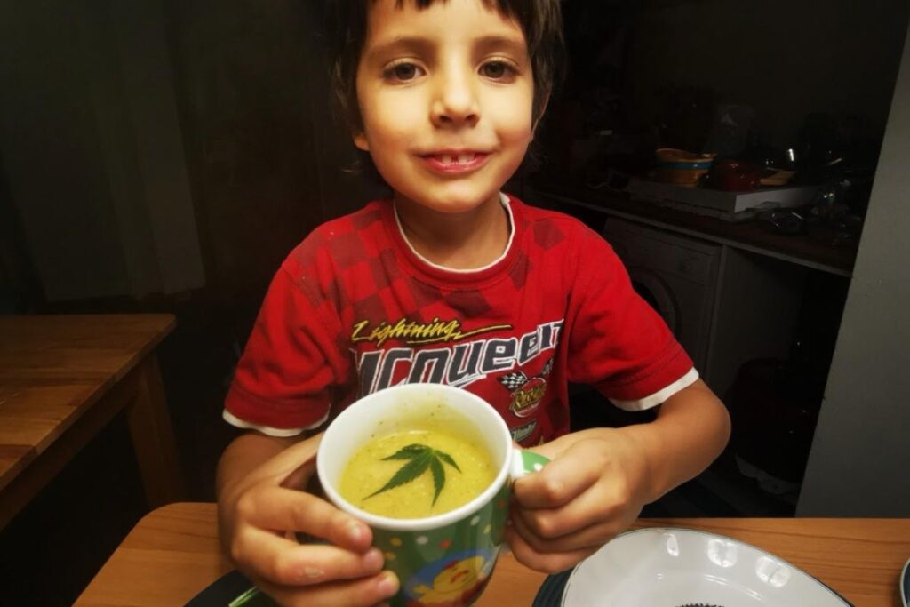 笑顔の男の子が両手で緑色のマグカップを持ち、黄色いスープ（表面に大麻の葉が浮いている）を飲もうとしている様子