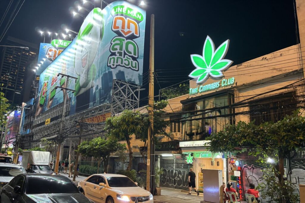 夜のバンコクの街並み。大きな緑の大麻の葉のネオンサインが輝く『Thai Cannabis Club』と、Line Manの巨大広告看板が目立つ繁華街の夜景