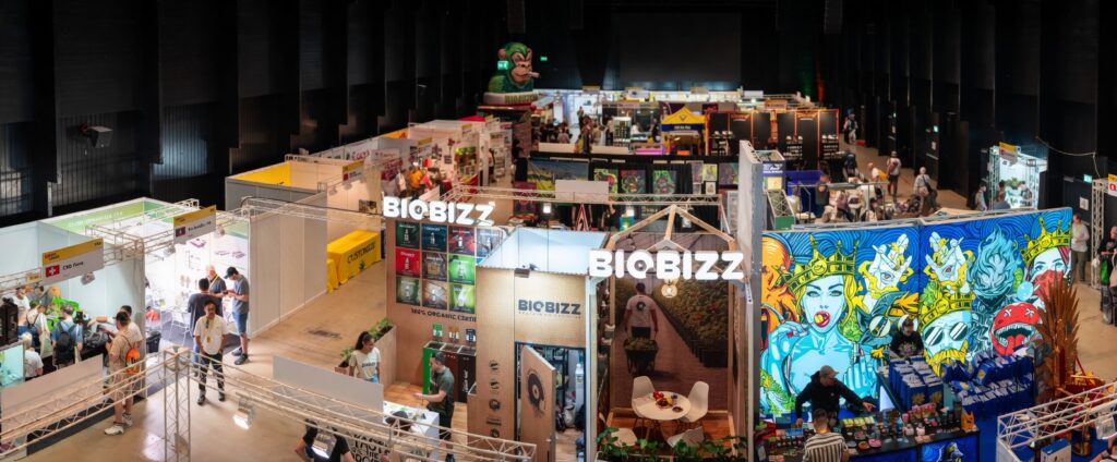BIOBIZZブースを中心とした大規模な国際ヘンプ・CBD展示会の会場風景。カラフルなアートウォールや多数の出展ブース、来場者で賑わう活気あるイベントホール（Canna Tradeや類似の国際展示会）の俯瞰写真。