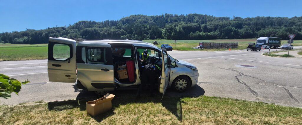 Canna Trade 会場に向かう道中の銀色バン。道路脇に停車し、ドアを開けて荷物（スーツケースや段ボール）を積み下ろし中の人々。晴れた青空の下、森と田園風景が広がるチューリッヒ近郊の道路風景。