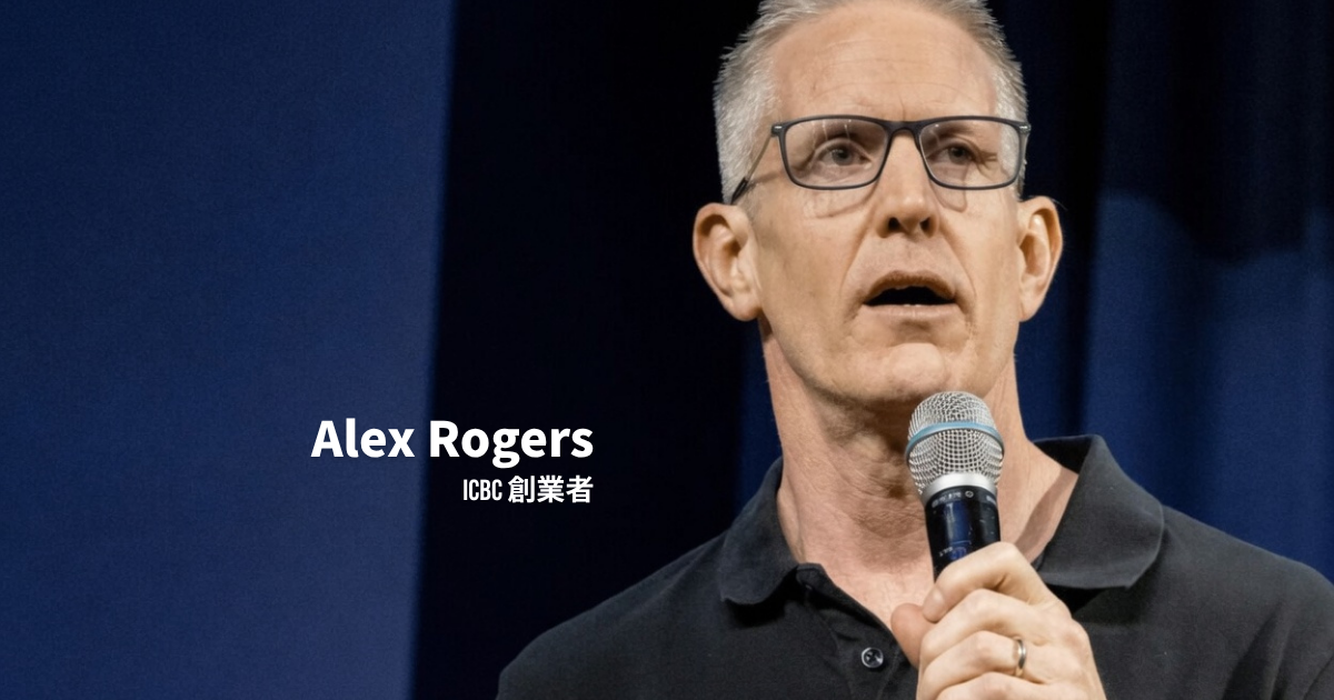 眼鏡をかけた灰色の短髪のICBC創業者、Alex Rogersが、黒いポロシャツを着てステージ上でマイクを持ちながら話している