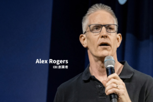 眼鏡をかけた灰色の短髪のICBC創業者、Alex Rogersが、黒いポロシャツを着てステージ上でマイクを持ちながら話している