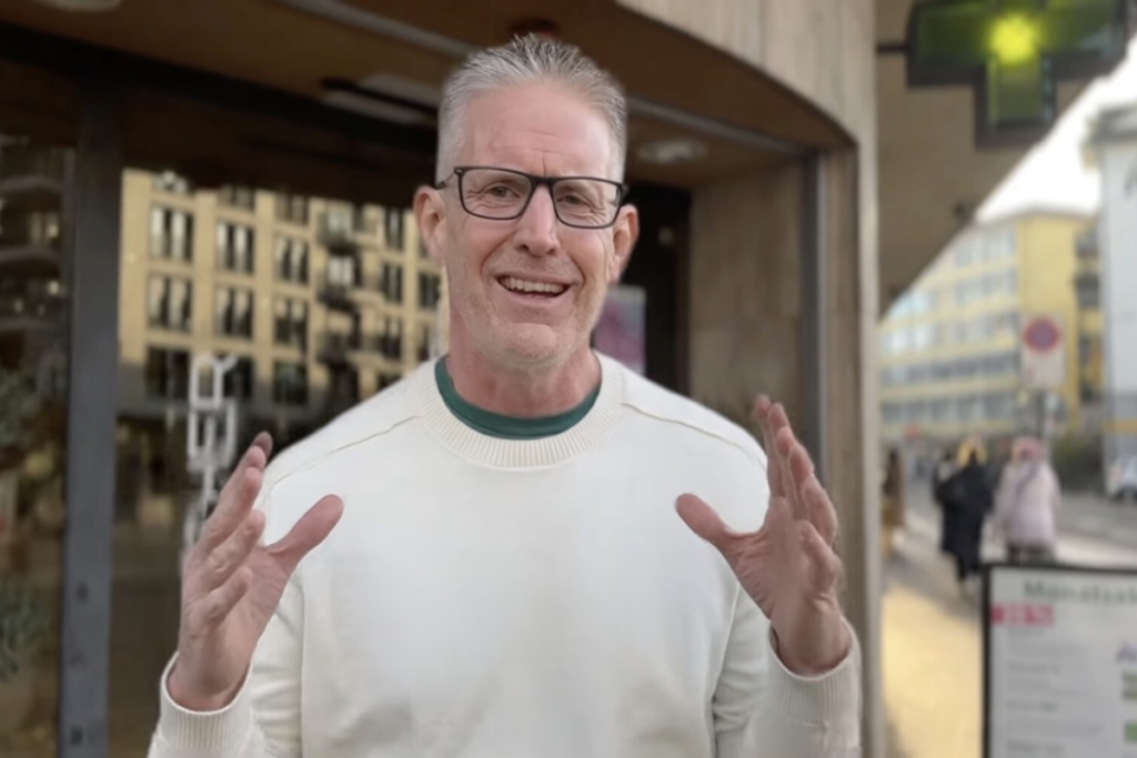 ICBC CEO Alex Rogersがスイス・チューリッヒの街中で白いセーター姿で両手を広げて挨拶する笑顔のポートレート。背景にチューリッヒの街並み