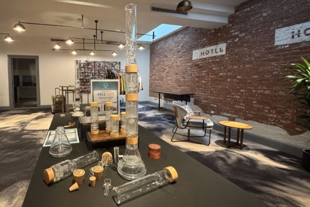 黒いテーブルの上にさまざまなガラス製ボングやアクセサリーが並んだ店舗内の写真。中央に木製リング付きの背の高いボングが立ち、背景にはレンガ壁、椅子、照明が見える。