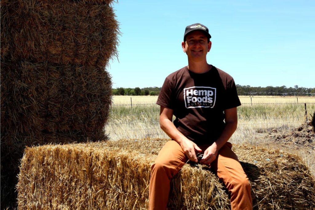 Hemp Foods AustraliaのTシャツとキャップを着た男性が、農場の干し草の山に座って笑顔でカメラを見つめている。背景に青空と広大な畑が広がる爽やかな屋外写真。