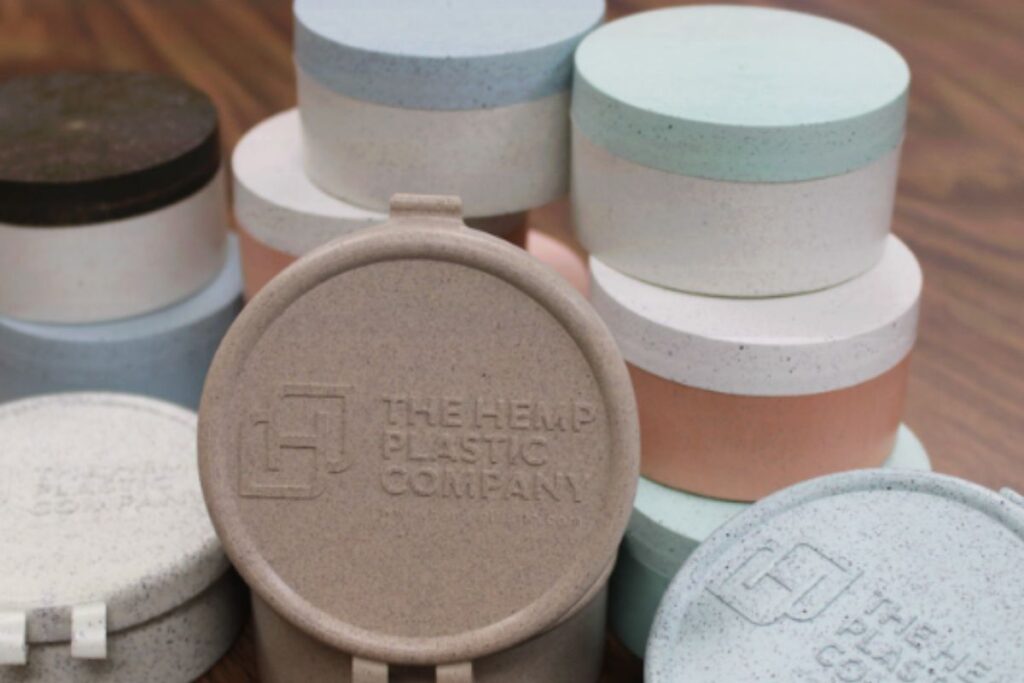 The Hemp Plastic Companyのヘンププラスチック製容器の蓋。ベージュ・ライトブルー・ミントグリーン・サーモンピンクなどの柔らかい色合いの丸い蓋が木の上で積み重ねられているクローズアップ写真。中央にロゴと文字が刻印されている。