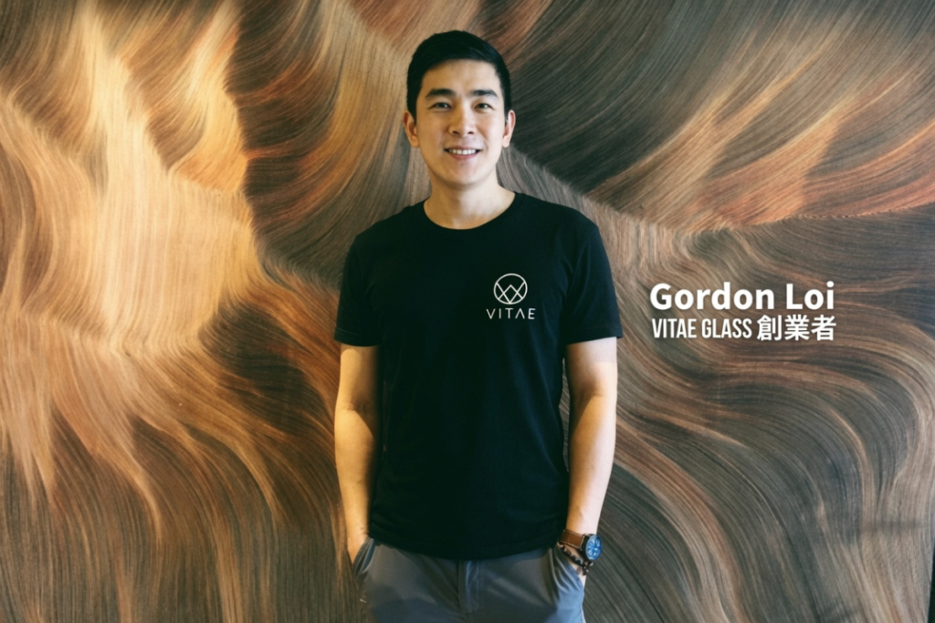 黒いTシャツにVITAEロゴを着用したGordon Loi氏が、流れるような美しい木目模様の背景の前で微笑んでいる写真。右側に「Gordon Loi VITAE GLASS 創業者