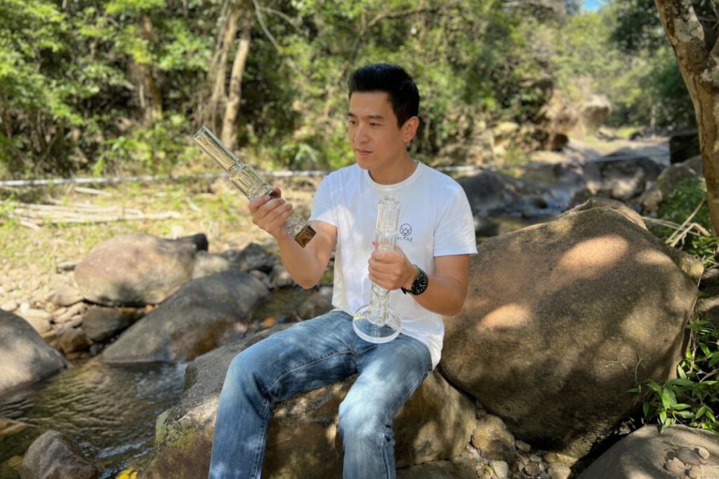 緑豊かな森の渓流沿いの大きな岩の上に座り、白いTシャツとジーンズ姿の男性が2つの透明なガラス製品を手に持って眺めている自然の中での写真