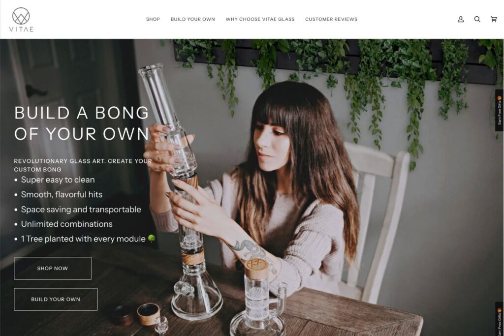 Vitae Glass公式サイトのヒーロー画像。暗い髪の女性がベージュのセーターを着て木製テーブルに座り、木目調パーツ付きのモジュール式クリアガラスボングを組み立てている様子。背景にハンギンググリーン、左側に「BUILD A BONG OF YOUR OWN」のキャッチコピーと特徴リスト、SHOP NOWとBUILD YOUR OWNのボタン