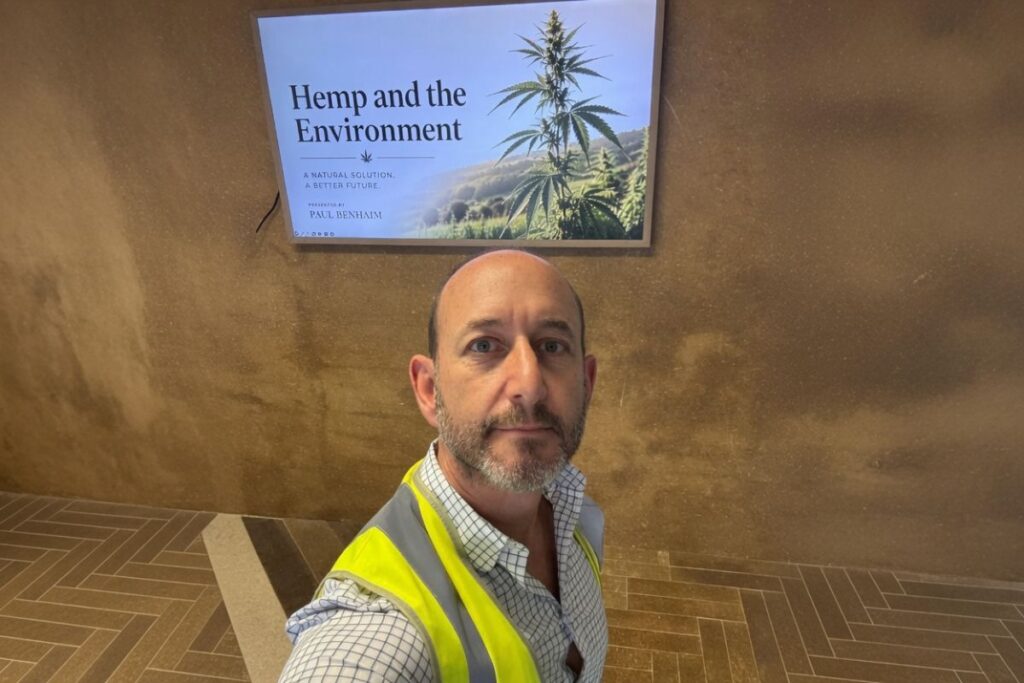 Paul BenhaimがHemp and the Environmentのポスターの前でセルフィーを撮っている写真。黄色の安全ベストを着用し、背景に大麻植物の大きなポスターが掛かっている。