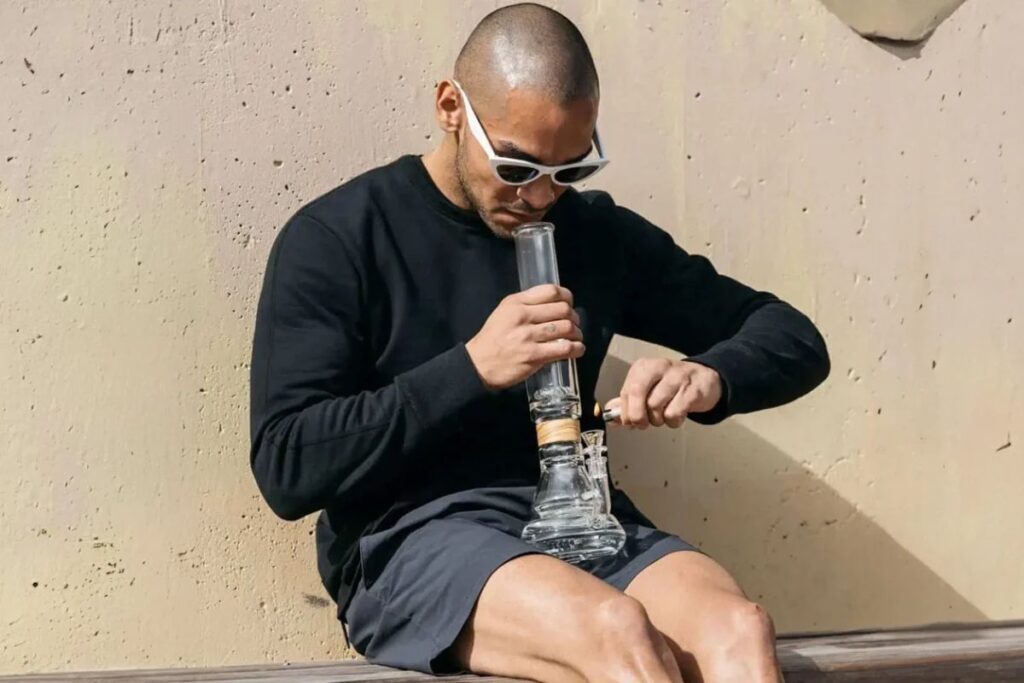 屋外でベンチに座り、ライターで火をつけながらガラス製のボングを使用する男性。