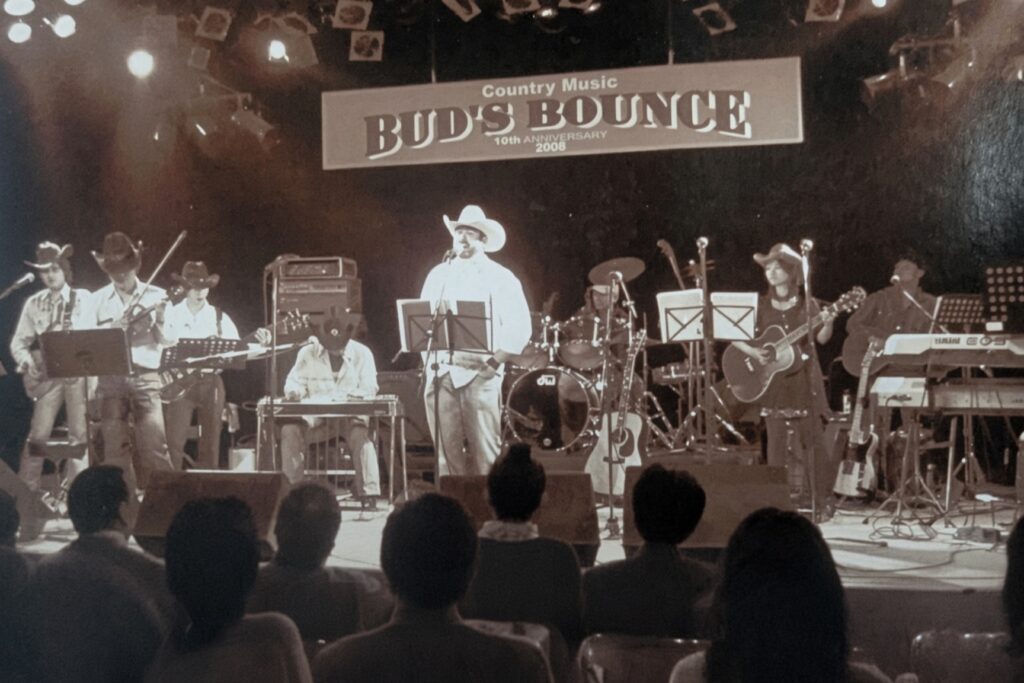 2008年開催のカントリーミュージック「BUD'S BOUNCE」10周年記念ライブのステージ写真。白いカウボーイハットをかぶったボーカリストを中心に、バイオリン、スチールギター、ドラム、アコースティックギター、キーボードを演奏するバンドメンバーが並び、前方には観客のシルエットが見えるモノクロ風のライブ写真。