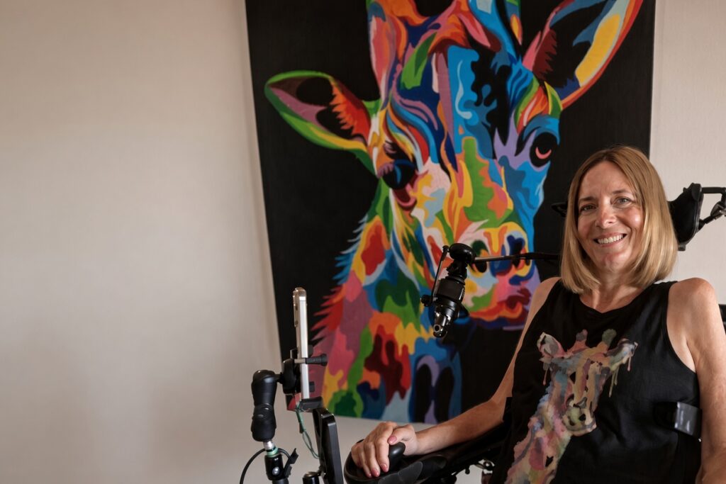 車椅子に座り、カラフルなキリンの絵画の前で微笑む金髪の女性。黒いキリン柄のタンクトップを着て、マイクアームとスマートフォンホルダーが付いた電動車椅子に座っている室内のポートレート写真。