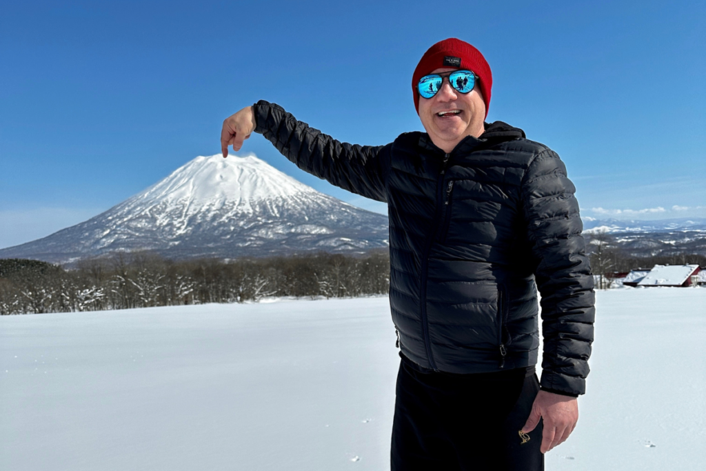 雪に覆われた富士山を背景に、赤いニット帽と青いミラーサングラスをかけた男性が山を指差して微笑んでいる冬の風景