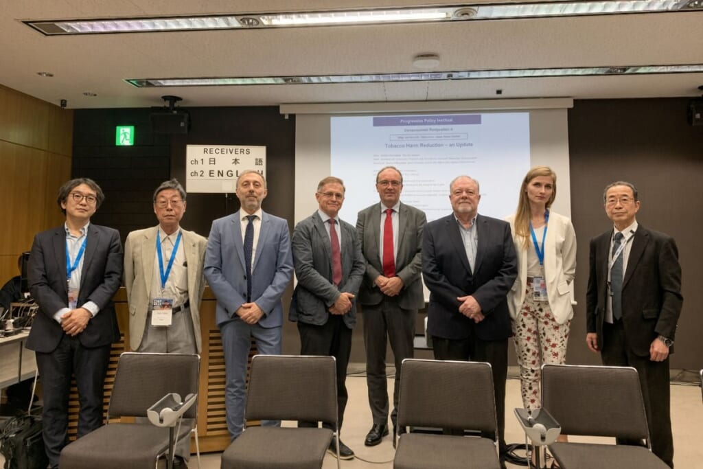 会議室で並んで立つ8人の男女。後ろに「Tobacco Harm Reduction – an Update」と書かれた投影スクリーンと「RECEIVERS」の看板がある。